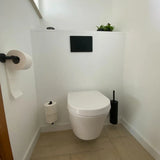 Een minimalistische badkamer met een wandcloset, twee wc-papierrollen op houders, een Corrado Black toiletset van yessential.nl (inclusief een zwarte wc-borstel), een plantje, een spoelpaneel op een witte muur en een lichtgekleurde tegelvloer.