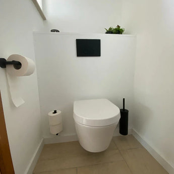Een minimalistische badkamer is voorzien van een wit wandtoilet, Tino Black spoelpaneel van yessential.nl, zwarte toiletaccessoires waaronder een toiletrolhouder zwart en borstel, twee toiletpapierrollen op houders en een klein groen plantje erboven.