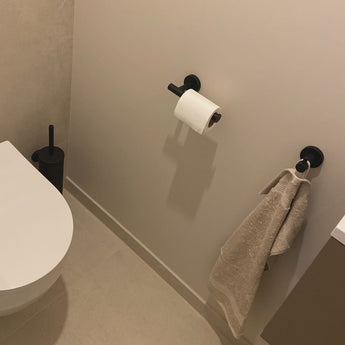Een minimale badkamerhoek met de yessential.nl Ferro Black WC set: een wit toilet, zwarte toiletborstelgarnituur, plus bijpassende handdoekring en papierhouder, allemaal gestyled tegen lichte muren en vloer.