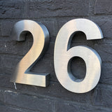 De Angelo RVS 20cm van yessential.nl is een groot metalen huisnummer 26, gemonteerd op een donkere bakstenen muur en werpt zachte schaduwen naar links voor een stijlvolle moderne look.