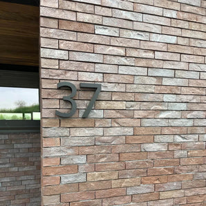 Het Carlo Black 20cm huisnummer van yessential.nl, met een strak 3D ontwerp, is gemonteerd op een lichtbruine bakstenen muur naast het raam met houten kozijnen van een modern gebouw.
