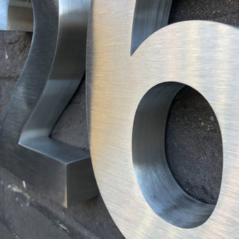 Close-up van het Angelo RVS 20cm huisnummer van yessential.nl, met zijn geborstelde metalen afwerking en 3D textuur op een donkere bakstenen muur.