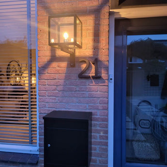 Een verlichte buitenlantaarn schijnt boven een zwarte brievenbus en een strak yessential.nl Carlo Black 20cm huisnummer, gemonteerd op de bakstenen muur naast een raam met jaloezieën en een deel van een blauwe deur.