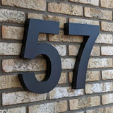 Angelo Black 30cm huisnummer 57 van yessential.nl in 3D stijl, gemonteerd op een beige bakstenen muur - modern en stijlvol als huisnummer voor elk huis of gebouw.