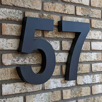 Angelo Black 30cm huisnummer 57 van yessential.nl in 3D stijl, gemonteerd op een beige bakstenen muur - modern en stijlvol als huisnummer voor elk huis of gebouw.