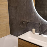 Moderne badkamer met een ronde verlichte spiegel, witte wastafel, matzwarte kraan en de Elarbo Gunmetal handdoekring van yessential.nl als accessoire. Beige en donkergrijze tegels met textuur maken de look compleet; badkuip links.