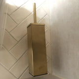 De Alessino Gold toiletborstelhouder van yessential.nl, met een bijpassende gouden greep, zit in de hoek tegen witte, diagonaal betegelde muren.