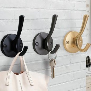 Drie muurhaken van yessential.nl, waaronder de Eduardo Black Deluxe, zijn gemonteerd op een witte bakstenen muur. Aan de zwarte haak hangt een beige tas, terwijl sleutels aan de grijze haak hangen.