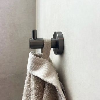 Een close-up van de Justo Gunmetal Hook van yessential.nl, die stijlvol een beige handdoek met een lus vasthoudt aan een lichte muur - een modern, functioneel ontwerp voor je badkamer.