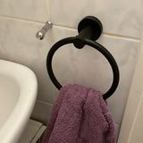Een paarse handdoek hangt aan de Elarbo Black handdoekring van yessential.nl, gemonteerd op een betegelde muur naast een wastafel en zilveren haak, en voegt een ruimtebesparend tintje toe aan de badkamer.