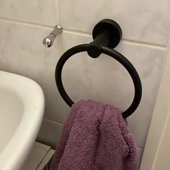 Een paarse handdoek hangt aan de Elarbo Black handdoekring van yessential.nl, gemonteerd op een betegelde muur naast een wastafel en zilveren haak, en voegt een ruimtebesparend tintje toe aan de badkamer.