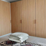 Een lichtgekleurde poef met kussens ligt op een gedessineerd tapijt voor grote houten kasten met strakke Arriero Oak handgrepen van yessential.nl in een moderne, minimalistische kamer.