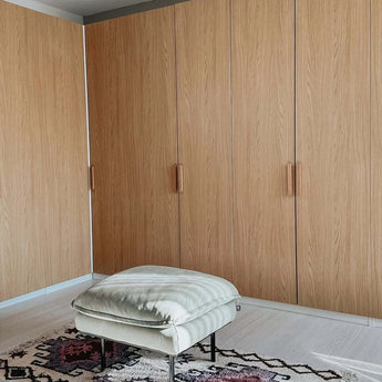 Een lichtgekleurde poef met kussens ligt op een gedessineerd tapijt voor grote houten kasten met strakke Arriero Oak handgrepen van yessential.nl in een moderne, minimalistische kamer.