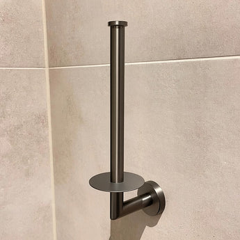 De Roberto Gunmetal toiletrollen houder van yessential.nl heeft een strakke, metallic uitstraling met een minimalistische verticale paal en platte ronde voet, en is afgebeeld gemonteerd op een beige betegelde muur.