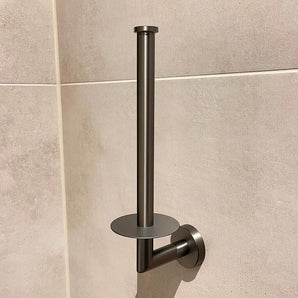De Roberto Gunmetal toiletrollen houder van yessential.nl heeft een strakke, metallic uitstraling met een minimalistische verticale paal en platte ronde voet, en is afgebeeld gemonteerd op een beige betegelde muur.