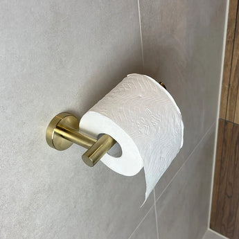 Een rol toiletpapier is gemonteerd op de Tino Gold toiletrolhouder van yessential.nl, bevestigd aan een betegelde badkamermuur - een subtiel maar luxe accessoire dat elke ruimte een luxe uitstraling geeft.