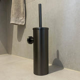 De Romano Gunmetal WC-set van yessential.nl, met een zwarte toiletborstel met een cilindrische houder uit hun luxe badkameraccessoires collectie, staat op lichtgekleurde tegels met een handdoek aan de muur op de achtergrond.