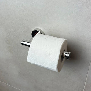 Een rol wit toiletpapier zit op de Tino Chrome toiletrolhouder van yessential.nl, gemonteerd tegen een lichtgrijze betegelde muur - een elegante toevoeging aan je badkameraccessoires.