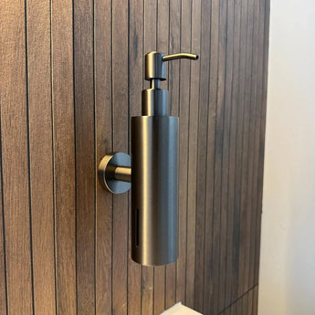 De Alberto Gunmetal zeeppomp van yessential.nl is een stijlvolle, cilindrische zeeppompje muur in gunmetal, gemonteerd op een badkamerwand met verticale houten panelen en voorzien van een strak pompje bovenop voor gemakkelijk gebruik.
