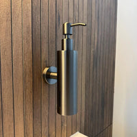 De Alberto Gunmetal zeeppomp van yessential.nl is een stijlvolle, cilindrische zeeppompje muur in gunmetal, gemonteerd op een badkamerwand met verticale houten panelen en voorzien van een strak pompje bovenop voor gemakkelijk gebruik.