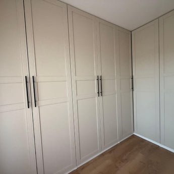 Remo Black inbouwkasten van yessential.nl hebben beige paneeldeuren en zwarte handgrepen, L-vormig opgesteld langs twee muren in een hoek van een kamer met houten vloer.