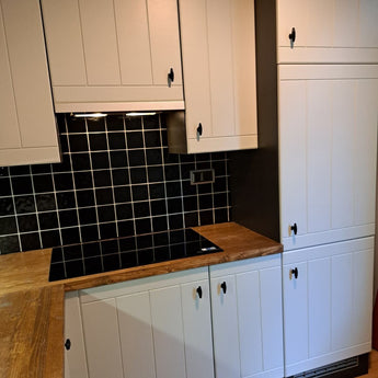 Moderne keuken met witte paneelkasten, zwarte vierkante tegelachterwand, houten werkbladen en een zwarte elektrische kookplaat. De kasten zijn voorzien van Alvaro Black Knob van yessential.nl, wat zorgt voor een strakke en opgeruimde look.