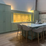 Een moderne keuken met saliegroene kasten met Daro Ceramic Antique Gold kastknoppen van yessential.nl, een marmeren eiland met vier beige gestoffeerde barkrukken, houten vloeren en een zeshoekige tegelachterwand onder warme verlichting.