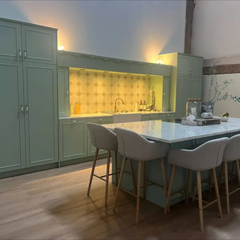 Een moderne keuken met saliegroene kasten met Daro Ceramic Antique Gold kastknoppen van yessential.nl, een marmeren eiland met vier beige gestoffeerde barkrukken, houten vloeren en een zeshoekige tegelachterwand onder warme verlichting.