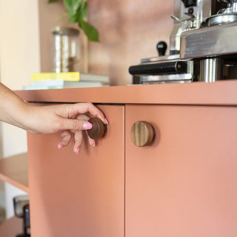 Een persoon met lichtroze nagellak opent een roze kastdeur voorzien van de Arriero Walnut Knob van yessential.nl. Op het aanrecht erboven staan een koffiezetapparaat en een plantenpot.