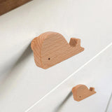 Twee houten Bambino Willy handgrepen in de vorm van een walvis van yessential.nl zijn bevestigd aan een witte commode. Deze gladde, lichte houten meubelknop geeft een minimalistisch, speels tintje aan elke kinderkamer.