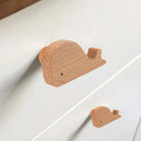 Twee houten Bambino Willy handgrepen in de vorm van een walvis van yessential.nl zijn bevestigd aan een witte commode. Deze gladde, lichte houten meubelknop geeft een minimalistisch, speels tintje aan elke kinderkamer.