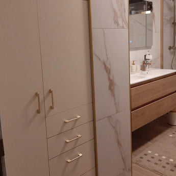 Creëer een moderne badkamer met Ramiro Gold Deluxe tegels met marmereffect van yessential.nl, een hoge kast met gouden handgrepen, een houten badmeubel en witte wastafel, een spiegel erboven en een vloerkleed met patroon voor een elegante afwerking.