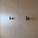 Twee Rodrigo Gunmetal haken van yessential.nl, gelijkmatig gemonteerd op een beige betegelde muur en zacht verlicht door natuurlijk licht, voegen een strak accent toe aan je badkamer.