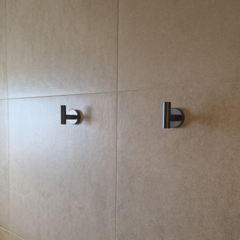 Twee Rodrigo Gunmetal haken van yessential.nl, gelijkmatig gemonteerd op een beige betegelde muur en zacht verlicht door natuurlijk licht, voegen een strak accent toe aan je badkamer.