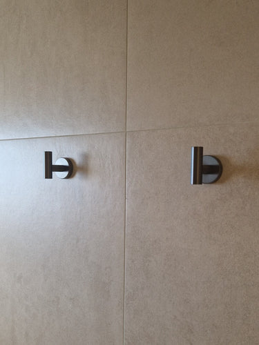 Twee Rodrigo Gunmetal haken van yessential.nl, gelijkmatig gemonteerd op een beige betegelde muur en zacht verlicht door natuurlijk licht, voegen een strak accent toe aan je badkamer.