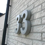 Grote, grijze Enrico beton 30cm huisnummers van yessential.nl zijn gemonteerd op een lichte bakstenen muur. De foto, die onder een hoek is genomen, benadrukt de 3D-dikte en werpt opvallende schaduwen op het oppervlak.