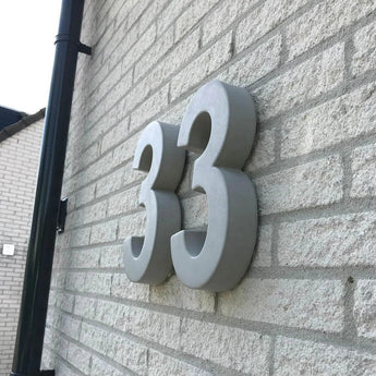 Grote, grijze Enrico beton 30cm huisnummers van yessential.nl zijn gemonteerd op een lichte bakstenen muur. De foto, die onder een hoek is genomen, benadrukt de 3D-dikte en werpt opvallende schaduwen op het oppervlak.