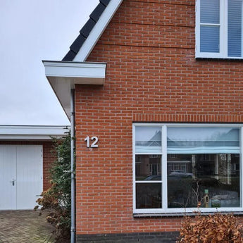 De foto toont de hoek van een rood bakstenen huis met een wit omlijst raam, een Enrico beton 20cm huisnummer 12 van yessential.nl op de muur, een deel van een witte garagedeur en struiken op de voorgrond.