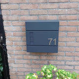 Het Animo RVS 7,6cm (geen boor) nummer van yessential.nl staat als "71" op een moderne donkergrijze brievenbus, gemonteerd op een bakstenen muur boven groene en gele planten.