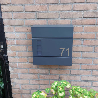 Het Animo RVS 7,6cm (geen boor) nummer van yessential.nl staat als "71" op een moderne donkergrijze brievenbus, gemonteerd op een bakstenen muur boven groene en gele planten.