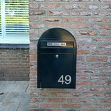 Een zwarte brievenbus met Animo RVS 7,6cm huisnummers van yessential.nl (geen boor) toont 49, gemonteerd op een bakstenen muur. Boven de brievenbus zitten twee NEE NEE stickers. Een raam met gesloten jaloezieën is gedeeltelijk zichtbaar aan de linkerkant.