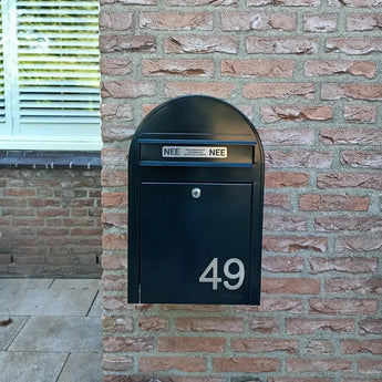 Een zwarte brievenbus met Animo RVS 7,6cm huisnummers van yessential.nl (geen boor) toont 49, gemonteerd op een bakstenen muur. Boven de brievenbus zitten twee NEE NEE stickers. Een raam met gesloten jaloezieën is gedeeltelijk zichtbaar aan de linkerkant.