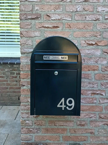 Een zwarte brievenbus met Animo RVS 7,6cm huisnummers van yessential.nl (geen boor) toont 49, gemonteerd op een bakstenen muur. Boven de brievenbus zitten twee NEE NEE stickers. Een raam met gesloten jaloezieën is gedeeltelijk zichtbaar aan de linkerkant.