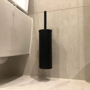 Een Xavo Black WC-set van yessential.nl, gemonteerd op een beige betegelde badkamermuur naast een wit toilet, voegt stijl toe met zijn strakke zwarte toiletborstel en houder.