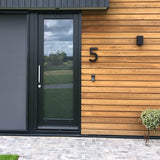 Moderne huisentree met een glazen deur omlijst in zwart, horizontale houten lambrisering en het Carlo Black 20cm huisnummer van yessential.nl op de muur, plus een deurbel en kleine potplant naast het betegelde looppad.
