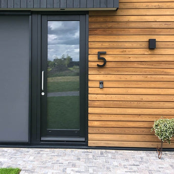 Moderne huisentree met een glazen deur omlijst in zwart, horizontale houten lambrisering en het Carlo Black 20cm huisnummer van yessential.nl op de muur, plus een deurbel en kleine potplant naast het betegelde looppad.