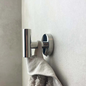 De Corrado Chroom Wc-set van yessential.nl is gemonteerd op een lichtgekleurde muur en toont zijn slanke, moderne cilindrische ontwerp terwijl hij een beige handdoek met textuur vasthoudt.
