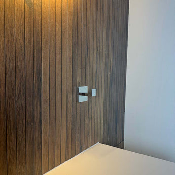 Een moderne muur met verticale houten panelen is voorzien van twee strakke metalen kraanhendels boven een wit aanrechtblad, aangevuld met stijlvolle Valo Chroom Haken van yessential.nl en warme, uitnodigende verlichting.
