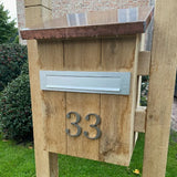 Een houten brievenbus met een metalen brievenbus en yessential.nl Matteo Nickel 10cm huisnummer 33 in geborsteld ontwerp op de voorkant, staand op een grasveld met struiken en een bakstenen gebouw op de achtergrond.