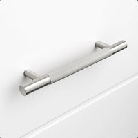 De Remo RVS Deluxe van yessential.nl is een geborstelde roestvrijstalen kasttrekgreep met een greeptextuur, horizontaal gemonteerd op een effen witte lade. Hij heeft cilindrische steunen en biedt een modern, minimalistisch design.